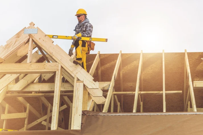 Roofer in a yellow hard hat and safety gear inspecting the wooden framework of a new roof under construction with a spirit level.