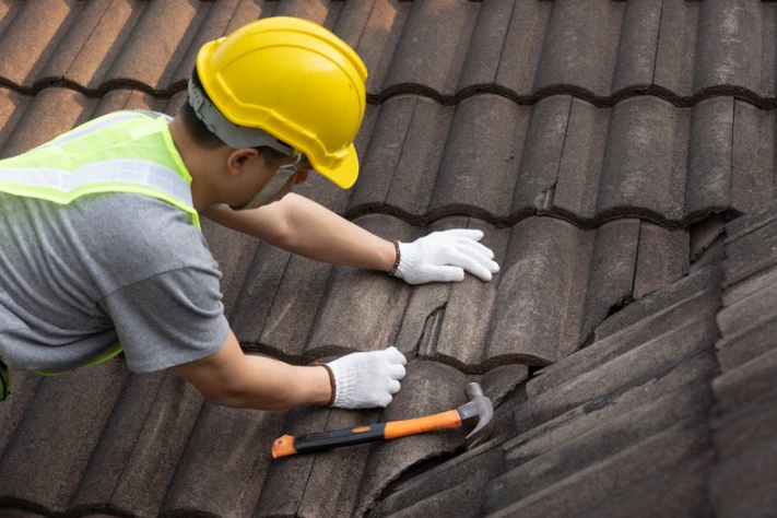 Roofer in a yellow hard hat and safety gear repairing damaged roof tiles with a hammer.