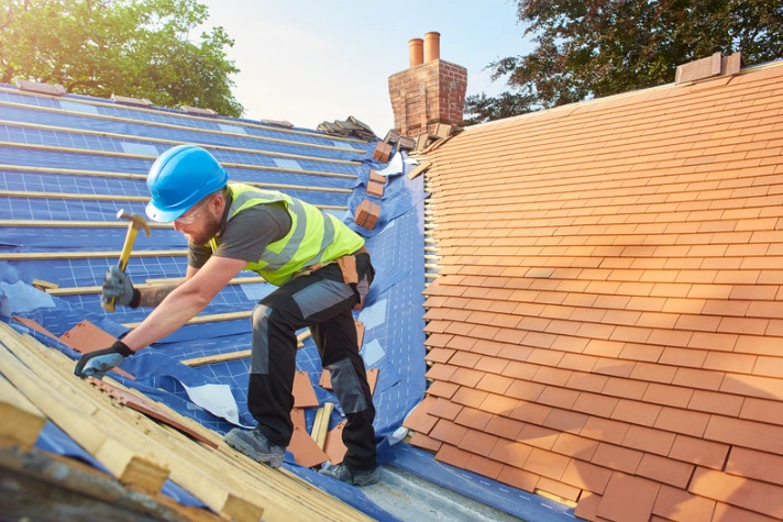 Roofer in a safety vest and hard hat installing new clay tiles on a residential roof during a roof replacement project.