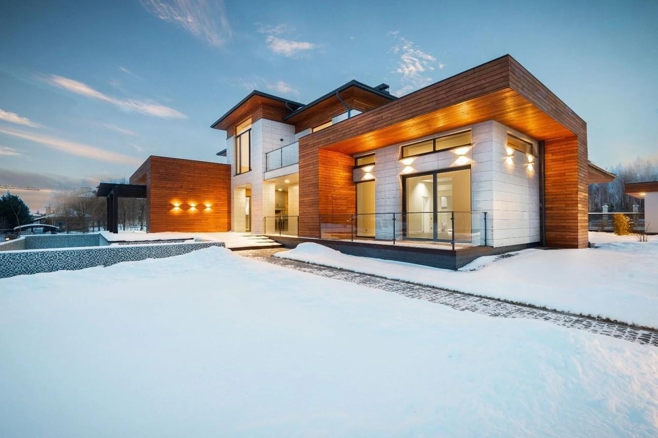 A modern luxury home with wood and stone exterior, covered in snow at dusk, featuring a durable roof installed by Silverline Roofing Ltd